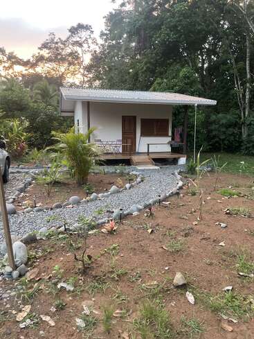 A small white house with a wooden porch sits among lush green trees. A gravel path, bordered by rocks, leads through a sparse garden.