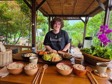 Une personne souriante est assise à une table en bois en plein air, entourée de plats sains et vibrants et de légumes frais, sous un gazebo rustique dans un décor de jardin verdoyant.
