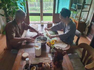 Deux personnes font de la pâte à pain ensemble, autour d'une table en bois, dans une pièce confortable et ensoleillée, avec des plantes, des ustensiles et une vue ouverte sur un jardin verdoyant.