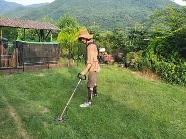 Un homme portant un chapeau de paille et des vêtements en lambeaux taille l'herbe avec un coupe-herbe dans un jardin verdoyant entouré de montagnes et d'arbres.