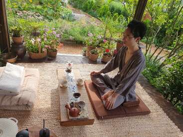 Une personne est assise sur un coussin, les jambes croisées, et profite d'une cérémonie du thé sereine dans un jardin paisible entouré de plantes en pot et de fleurs épanouies.