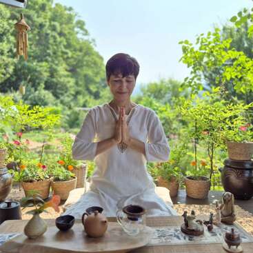 Uma mulher de branco medita pacificamente ao ar livre, com as mãos juntas, cercada por vasos de plantas, um jogo de chá e a natureza, exalando serenidade e calma à luz do dia.