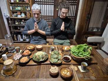 Two people sit at a wooden table with hands in prayer. Various Korean dishes, tea, and vegetables are arranged, creating a warm, traditional dining atmosphere.