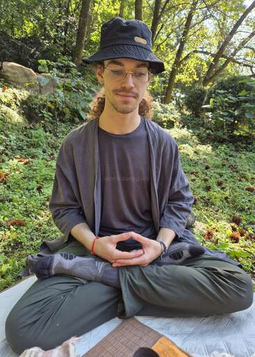 A person sits cross-legged on a blanket outdoors, meditates peacefully with hands in a mudra, wearing glasses, a black hat, and comfortable clothing. Lush greenery surrounds.