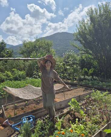 A smiling person wearing a straw hat stands in a lush vegetable garden, surrounded by greenery and flowers, with mountains and a bright blue sky behind.
