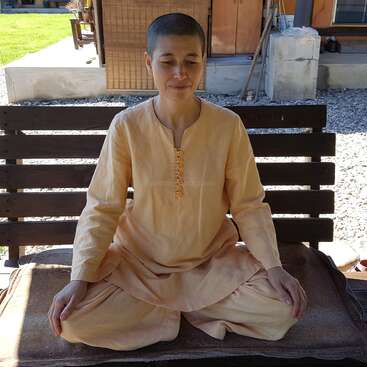 Uma pessoa com a cabeça raspada, vestindo roupas tradicionais laranja claro, senta-se de pernas cruzadas em um banco de madeira ao ar livre, meditando pacificamente sob a luz natural do dia.