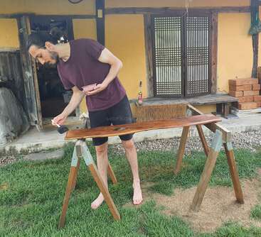 A man is barefoot, varnishing a wooden plank outdoors, standing on grass, in front of a rustic house with yellow walls, wooden beams, and traditional doors.