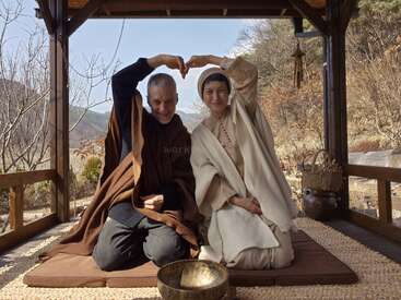 A joyful couple sits cross-legged outdoors, dressed in simple robes. Together, they make a heart shape with their arms, radiating peace and love in nature.