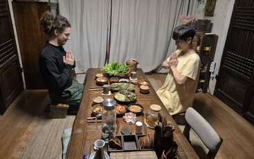 Two people sit across from each other at a wooden table, hands in prayer pose, about to share a meal of various traditional dishes in a cozy room.
