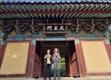 Two people stand respectfully with hands together beneath an ornate traditional Asian temple gate, featuring colorful wooden beams, detailed artwork, and a sign with Chinese characters.