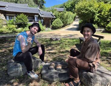 Two men sitting on rocks outdoors, enjoying a meal with chopsticks. Traditional Korean-style houses and lush greenery in the background, both smiling and relaxed.