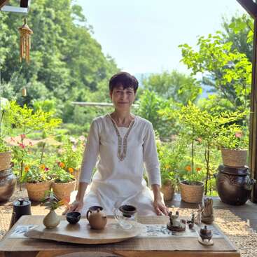 Eine Frau in einem weißen Kleid sitzt friedlich an einem Holztisch im Freien, umgeben von Topfpflanzen, Teeservice und Natur, und strahlt Gelassenheit und Ruhe aus.
