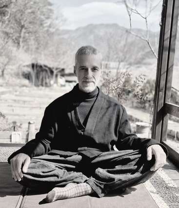 A man sits cross-legged on a mat, wearing traditional dark clothing. He is calm, with mountains and trees in the background, exuding peace and meditation.