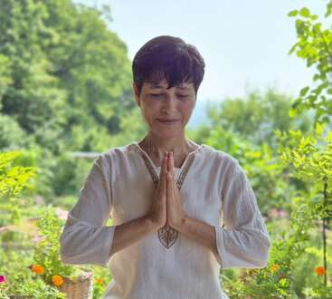 Una mujer está de pie al aire libre, con los ojos cerrados, las manos juntas en gesto de namaste, vestida con un ligero top blanco, rodeada de exuberantes plantas y flores verdes.