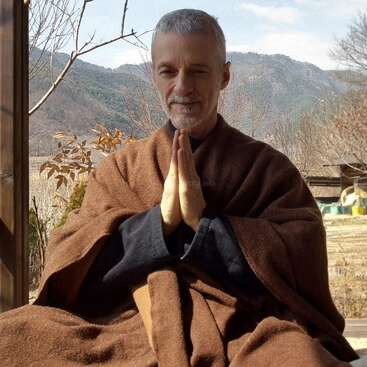 Un homme serein aux cheveux gris est assis dehors, enveloppé dans une robe marron, les mains jointes dans un geste de prière ou de méditation, avec les montagnes en arrière-plan.