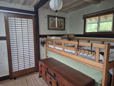Um quarto aconchegante com beliche de madeira, porta de correr japonesa, cômoda, quadros na parede, lanterna, janela e decoração rústica, com luz natural e tons terrosos.