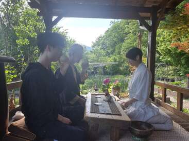 Tres personas se sientan juntas para celebrar una ceremonia tradicional del té al aire libre, rodeadas de exuberante vegetación y flores, creando un ambiente tranquilo y sereno bajo un pabellón de madera.