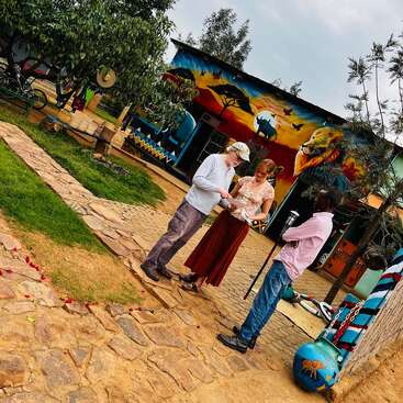La imagen muestra a tres personas en el exterior de un vibrante edificio con obras de arte de inspiración africana, rodeado de un camino de piedra y exuberante vegetación.