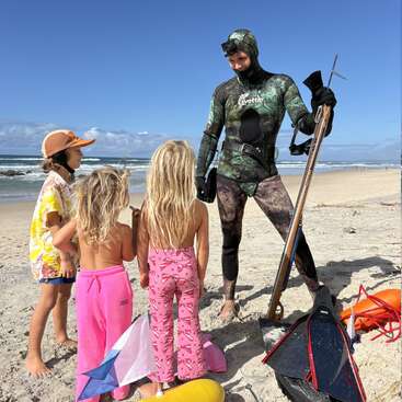 Un buceador con un traje de neopreno de camuflaje está de pie en una playa de arena, con un arpón en la mano, hablando con tres niños curiosos. Al fondo se ven un cielo despejado y las olas del mar.