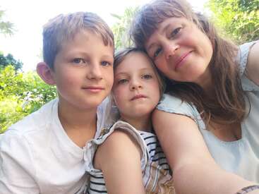 A woman and two children pose closely together, smiling outdoors with greenery in the background. The photo captures a sweet family moment in natural sunlight.