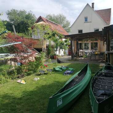 Das Bild zeigt eine ruhige Hinterhofszene mit zwei grünen Kanus im Gras, umgeben von einem Haus, Bäumen und einem wolkenverhangenen Himmel, der ein Gefühl der Ruhe ausstrahlt.