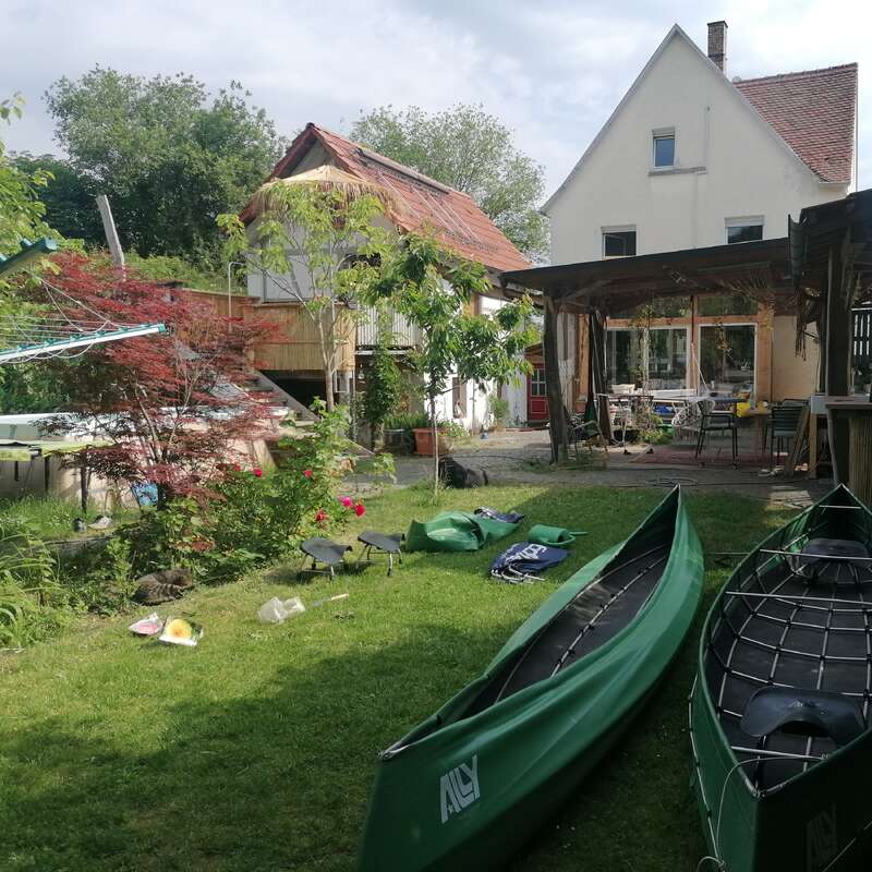 La imagen representa una serena escena de patio trasero con dos canoas verdes sobre la hierba, rodeadas de una casa, árboles y un cielo nublado, que desprenden una sensación de tranquilidad.