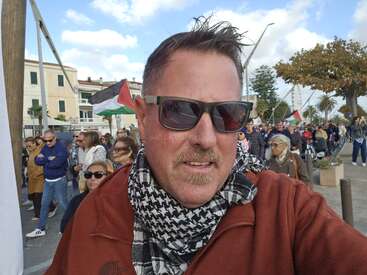 A man wearing sunglasses and a scarf takes a selfie at a street protest or march. People and Palestinian flags are visible in the background. It's daytime.