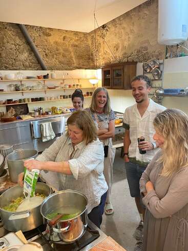 Five people gather in a cozy rustic kitchen, smiling and watching as one woman pours cream into a pot on the stove, creating a warm, communal atmosphere.