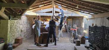Several people work inside a rustic building with wooden beams and a stone wall. One person stands on a ladder while others carry and stack roof tiles.