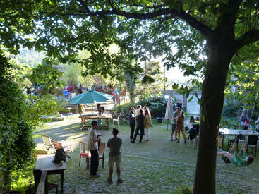 A group of people gather in a shaded, leafy garden, socializing around tables and chairs, some standing, others sitting or swinging, under a sunny sky.