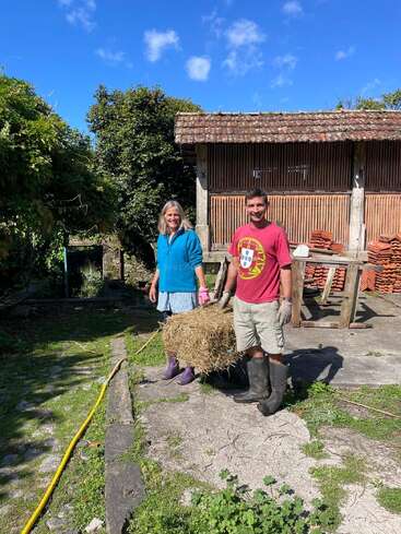 Two people, smiling, are carrying a bale of hay outdoors. The woman wears a blue jacket and purple boots. Behind them is a rustic wooden building and greenery.