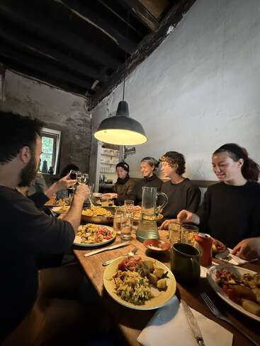 Five people sit around a rustic wooden table, sharing a hearty meal under warm lighting. Laughter and conversation fill the cozy, warmly lit, casual dining space.
