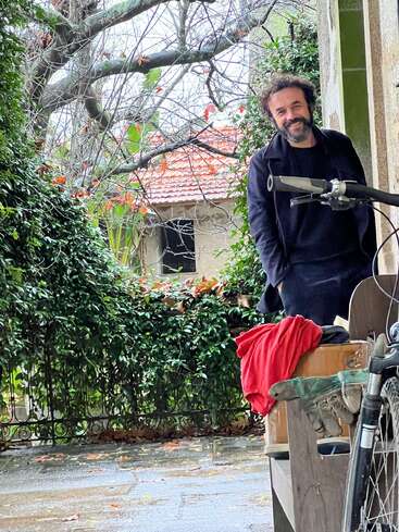 A man with curly hair and a beard smiles near a stone wall. A bicycle, red shirt, boots, and greenery surround him; a house is visible behind.