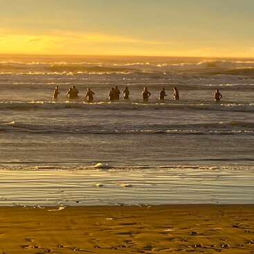 Eine Gruppe von Menschen steht bei Sonnenuntergang im Meer und hebt sich als Silhouetten vom goldenen Himmel ab. Sanfte Wellen rollen heran; im Vordergrund glitzert der nasse Sand.