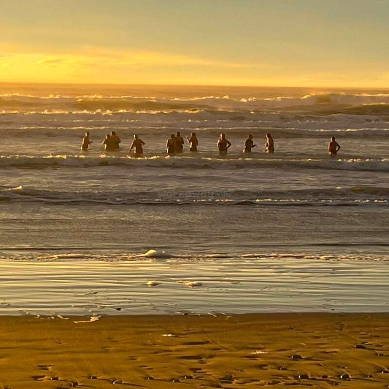 Um grupo de pessoas está na beira do mar ao pôr do sol, com suas silhuetas recortadas contra o céu dourado. Ondas suaves quebram na areia; a areia molhada brilha em primeiro plano.