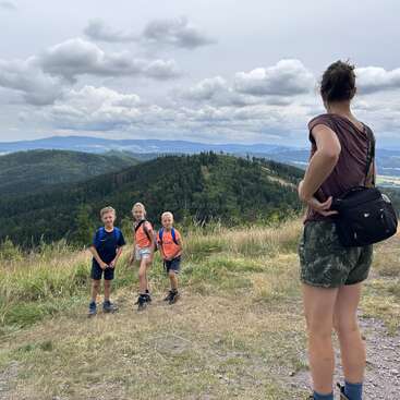 Une femme se tient sur un sentier de montagne herbeux, regardant trois enfants souriants avec des sacs à dos. Des collines, des forêts et des nuages spectaculaires forment l'arrière-plan.