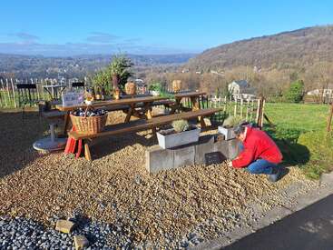 Jemand in einer roten Jacke kniet neben Pflanzkübeln in der Nähe eines Picknicktisches, der für Essen zum Mitnehmen gedeckt ist, und schaut auf eine malerische, hügelige Landschaft unter einem klaren blauen Himmel.