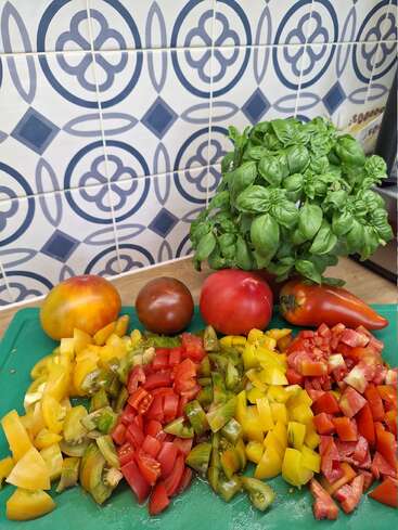 Bunte Tomatenwürfel verschiedener Sorten sind auf einem grünen Schneidebrett angeordnet, mit frischem Basilikum und ganzen Tomaten im Hintergrund, vor einem gemusterten Fliesenhintergrund.