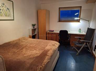 Ein gemütliches Schlafzimmer mit einem Bett, das mit einer braunen Decke zugedeckt ist, einem Kleiderschrank aus Holz, einem Schreibtisch, einem Bürostuhl, einer Leselampe, einer Topfpflanze und einem Fenster, durch das man den dunklen Himmel sehen kann.