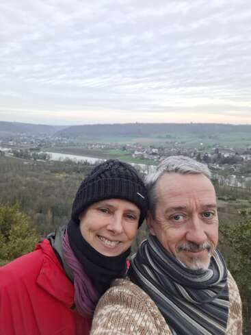 Ein lächelndes Paar, das mit Mützen und Schals warm angezogen ist, macht draußen ein Selfie mit einer coolen Kulisse aus Hügeln, einem Fluss, einem Dorf und einem bewölkten Himmel.