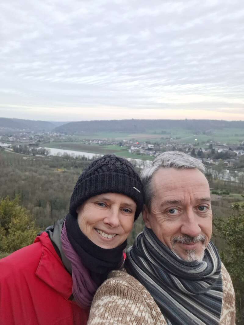 Ein lächelndes Paar, das mit Mützen und Schals warm angezogen ist, macht draußen ein Selfie mit einer coolen Kulisse aus Hügeln, einem Fluss, einem Dorf und einem bewölkten Himmel.