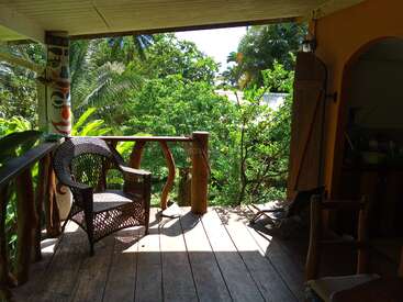 A cozy wooden porch with rustic railings, wicker chairs, and vibrant tropical greenery. Sunlight filters through, creating inviting shadows. A painted pillar adds artistic charm.