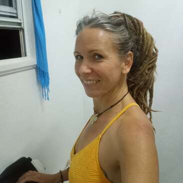 Uma mulher sorridente com longos dreadlocks e um top amarelo está em um ambiente interno. Há um lenço azul pendurado em uma janela aberta e paredes brancas ao redor dela.