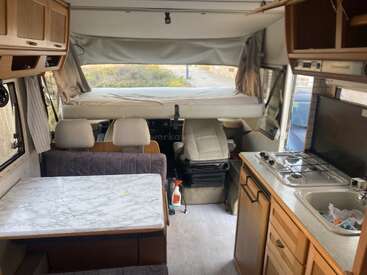 Cette image montre l'intérieur d'un camping-car avec une table, un coin cuisine, un comptoir, des armoires, des sièges et le poste de conduite à l'arrière-plan. Confortable et fonctionnel.