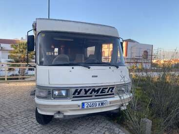 Un camping-car Hymer blanc immatriculé 2469 KWH est garé dans une rue pavée, à proximité de maisons et de feuillages, sous un ciel bleu clair.