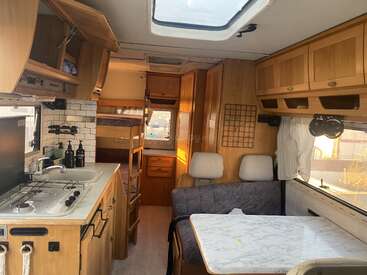 Cette image montre l'intérieur d'un camping-car confortable, avec une petite cuisine, des lits superposés, un coin salon avec une table et de nombreuses armoires de rangement en bois.