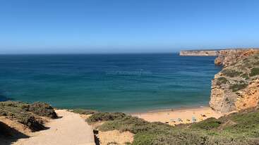 Un chemin pavé descend entre des buissons verts jusqu'à une plage de sable avec des parasols colorés. Les eaux claires et turquoises de l'océan rencontrent des falaises spectaculaires sous un ciel bleu sans nuage.