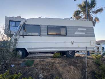 Un camping-car de collection est garé sur un terrain en terre battue, à proximité de bâtiments modernes et d'un palmier. Le ciel est dégagé, ce qui laisse présager une fin d'après-midi ensoleillée.