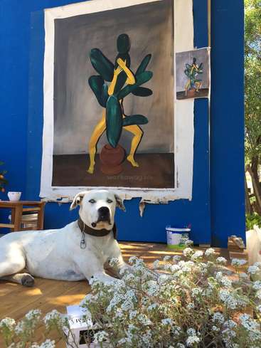 The image depicts a white dog lying on a wooden floor in front of a large painting of a person with a plant body, surrounded by flowers and a blue wall.