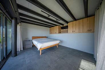 Cette chambre moderne et minimaliste comprend un lit en bois, une table de chevet, un grand carrelage, des armoires encastrées en bois, des poutres apparentes au plafond et de grandes fenêtres ornées de voilages.